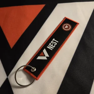 A close-up photo of a rectangular embroidered keychain featuring the NEST logo and lettering, measuring approximately 13 cm by 3 cm, with durable fabric and reinforced stitching.